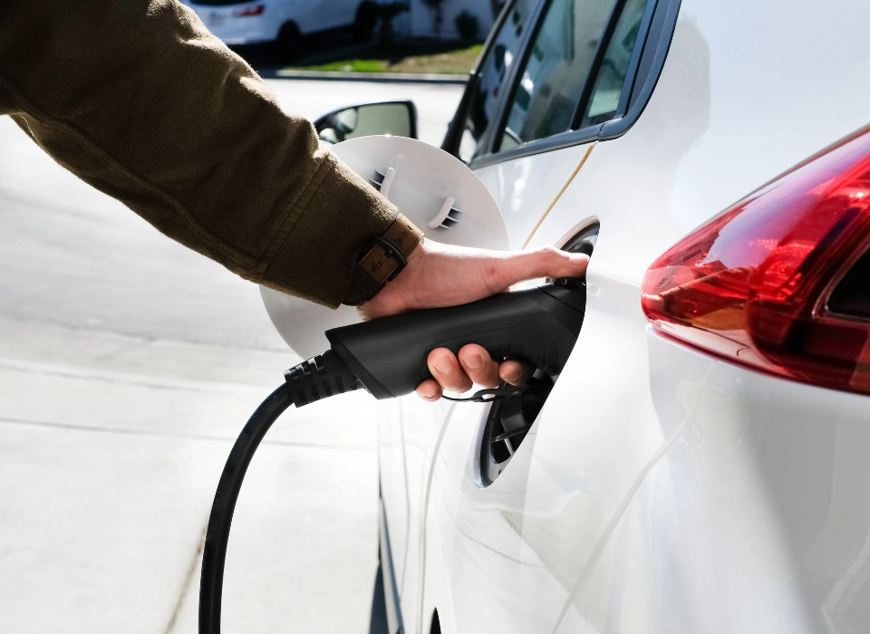 Persona conectando un cargador de auto eléctrico a un vehículo blanco en una estación de carga al aire libre.